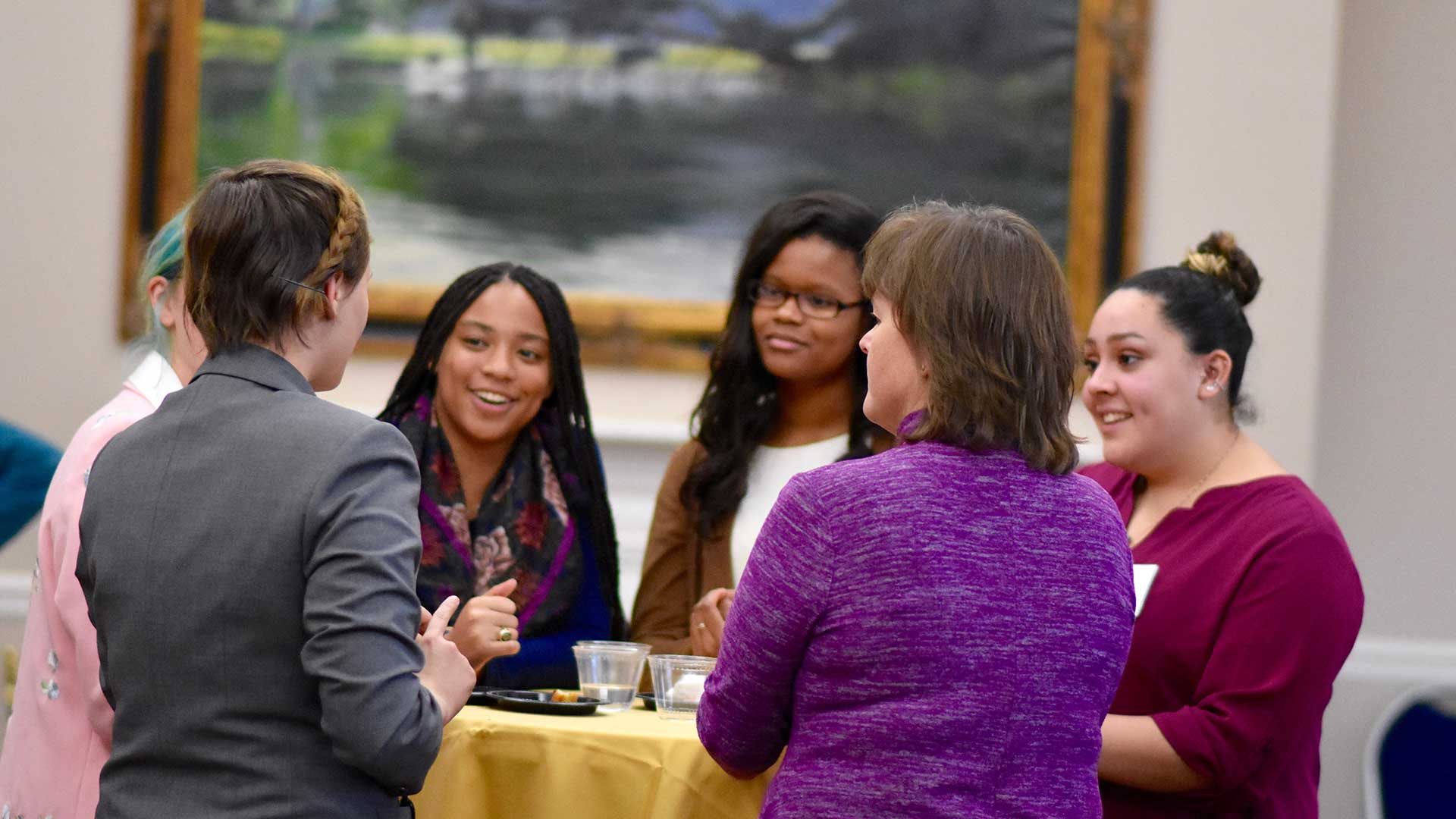Six women stand around a table having a conversation at an indoor event, with drinks and snacks on the table and a painting in the background.