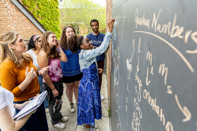 A group of students and a teacher gather outdoors around a large blackboard.