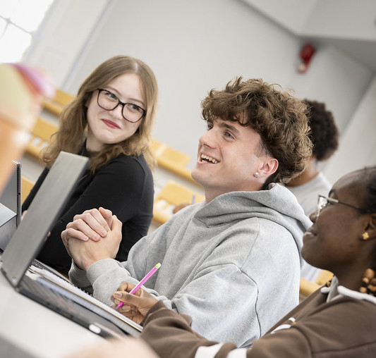 A group of students sit at desks with laptops in a classroom, engaging in conversation and taking notes.