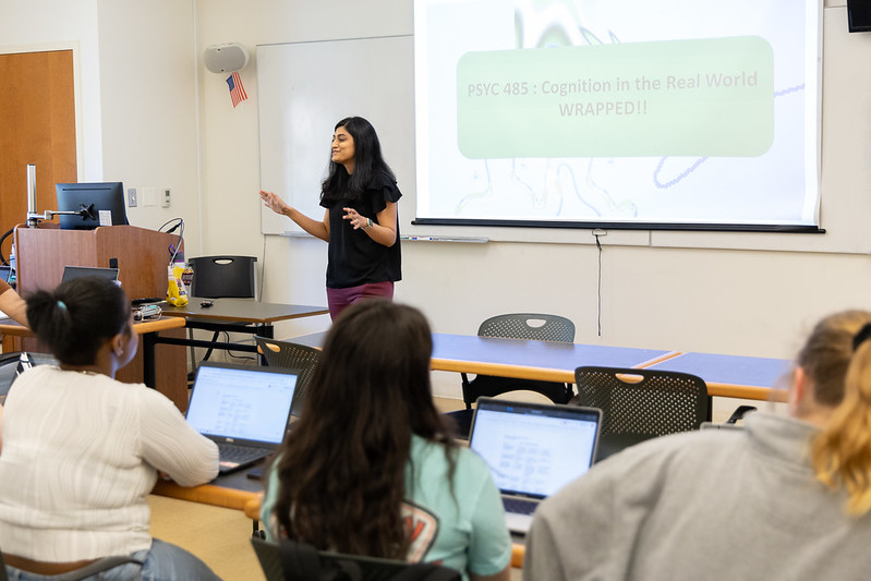 A woman stands at the front of a classroom presenting a slideshow titled "PSYC 485: Cognition in the Real World WRAPPED!" to students using laptops.