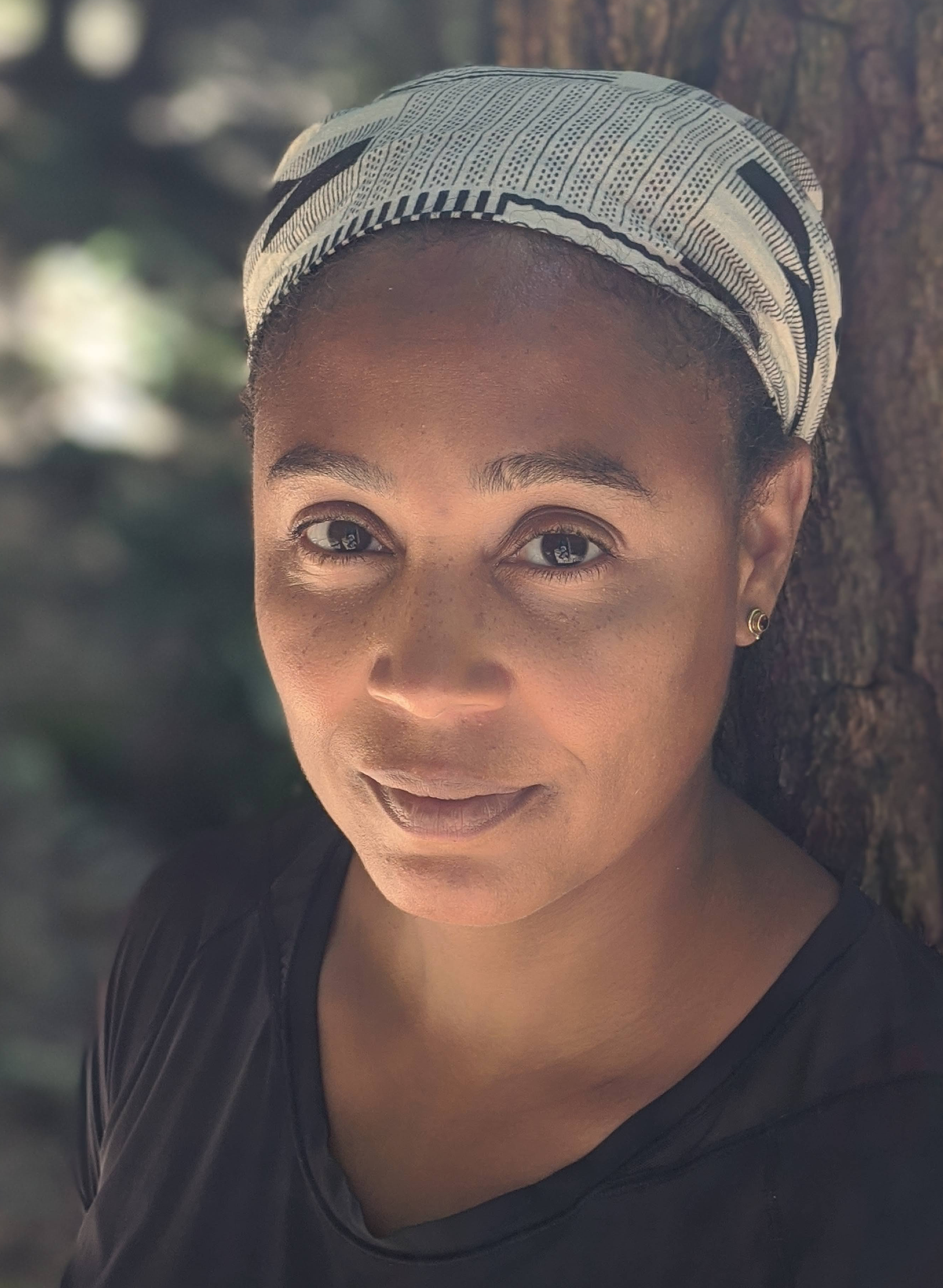 A woman wearing a patterned headscarf and black top stands outdoors, leaning against a tree, looking directly at the camera.